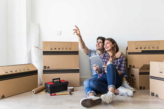 Young couple of man pointing up and woman holding tablet and smiling sitting on floor near boxes and tools