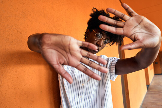 Adult African American female with eyes closed trying to stop negative impact while standing with arms raised against blurred vivid orange interior in corridor of modern building