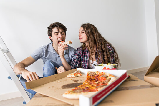 Cute young couple sitting on floor eating pizza at carton box near ladder and laughing