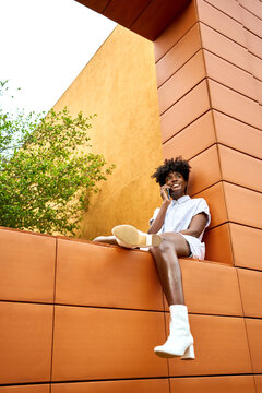 Low angle of young African American female in trendy white outfit smiling away and making phone call on street while sitting on wall contemporary building with bright orange and yellow walls