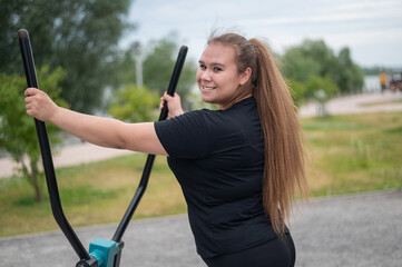 Beautiful smiling overweight young woman does fitness on an ellipsoid simulator outdoors. Fat girl...
