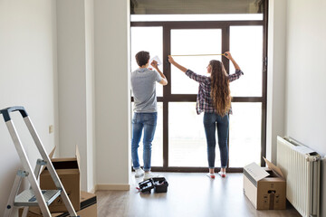 Young male and female measuring window with linear tape in apartment with carton boxes