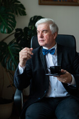 Confident elderly man in a suit and bow tie sits in a chair at his desk and drinks coffee. Portrait of a gray-haired businessman in an office.
