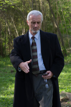 Well Dressed Businessman, Hand Extended But Looking Down At Smart Phone, Suit & Tie With Long Dark Blue Wool Overcoat, Three Quarter Length Portrait Stepping Toward Camera With Green Wooded Background