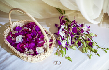 two silver wedding rings, a basket with white and purple petals of tropical flowers, a bouquet of white-purple orchids lie on a white bed. top view