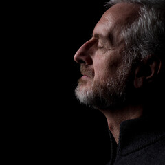 Middle aged man with gray hair and beard,  close up profile with hard shadows on black background with eyes closed with face and gaze slightly upward in thought, prayer, meditation or contemplation