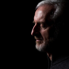Middle aged man with gray hair and beard,  close up profile with hard shadows on black background with eyes closed in thought, prayer, meditation or contemplation