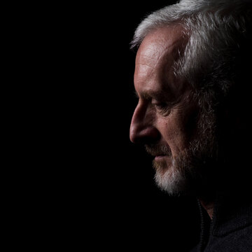 Middle aged man with gray hair and beard,  close up profile with hard shadows on black background with eyes open with face and gaze downward in thought, prayer, meditation or contemplation