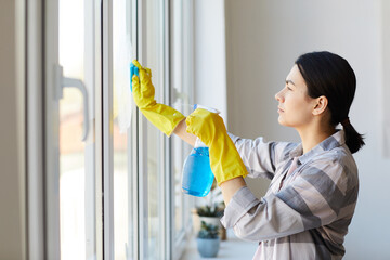 Young housekeeper in rubber gloves using cleanser spray and sponge and cleaning the window
