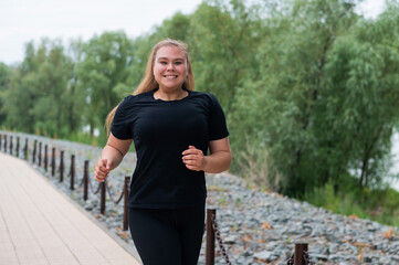 An obese young woman jogging outdoors. Fat beautiful smiling girl in a black tracksuit is engaged in fitness for weight loss on the waterfront. A woman runs on a summer day.