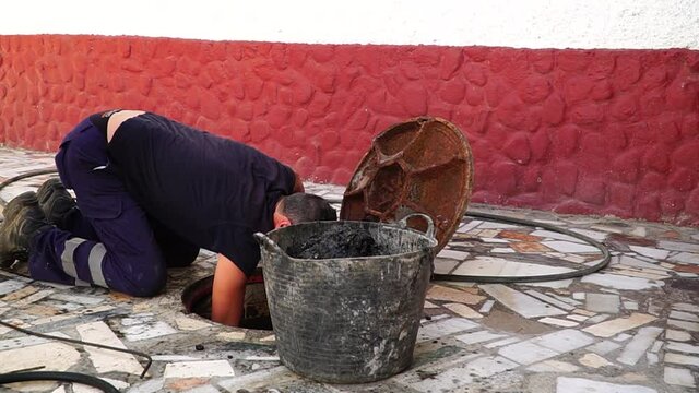 Worker cleans the drains hatch and removes dirt and debris from the sewer on the street. Repair of sewage or underground utilities, cleans drains. Plumber cleans the sewer.
