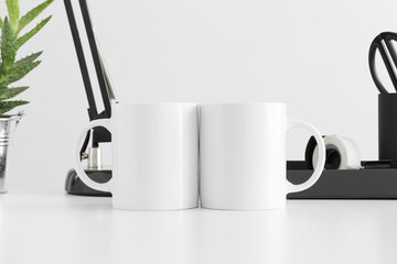 Two mugs mockup with a lamp, aloe vera plant and workspace accessories on a white table.