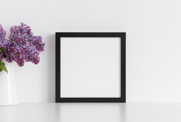 Black square frame mockup with a bouquet of lilac in a vase on a white table.