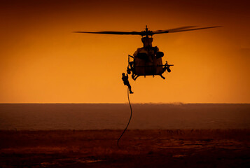 Shihoette of helicopter lowering a soldier by rope to the ground with ocean in view at sunset - obrazy, fototapety, plakaty