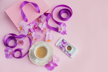 Flower overhead composition on light pink background top view. Cup of coffee, pink gift bag with purple ribbons, phone and pink orchid flowers.