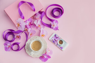 Obraz premium Flower overhead composition on light pink background top view. Cup of coffee, pink gift bag with purple ribbons, phone and pink orchid flowers.