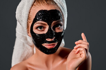 Portrait of a young attractive woman with a towel on her head with a cleansing black mask on her face isolated on a gray background.