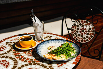 A breakfast consisting of an omelette with arugula or rocket salad, a yellow cup of black coffee, on a mosaic stone table of a cafe on a sunny day