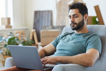 Young bearded man resting on armchair with laptop computer in the room at home