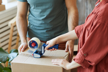 Close-up of young couple packing things in boxes together and using adhesive tape during moving in house