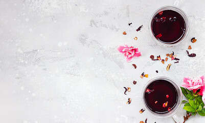 Two glass cups with red hot hibiscus tea on light grey concrete background with pink flowers and dry tea leaves. Top view, copy space.