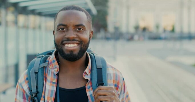Portrait of handsome young happy African American man with backpack smiling joyfully to camera outdoors. Close up of male tourist smiled face. At bus stop. Cheerful stylish guy traveller at street.