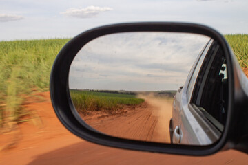 A rural dirt road and vast farmland reflected in the side mirror of a moving car on a sunny day,...