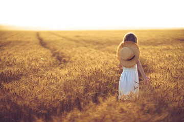 Fashion photo of a little girl in white dress and straw hat at the evening wheat field. Back view