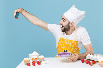 Excited young bearded male chef or cook baker man in apron white t-shirt toque chefs hat cooking at table isolated on blue background studio. Cooking food concept. Doing selfie shot on mobile phone.
