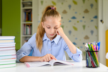 A tired girl sits with a book at the table and reads. Holds his head with his hand.