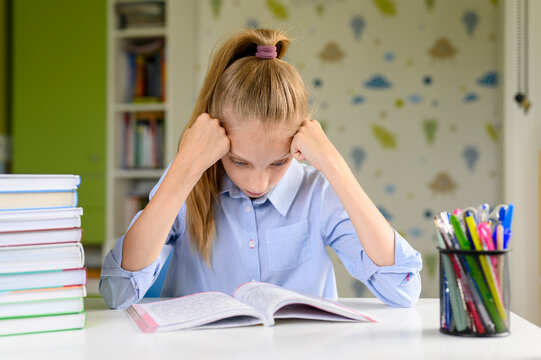 The schoolgirl in a bad mood rests her head on her hands and looks at the textbook. The girl is preparing for the exam.