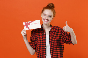Smiling young readhead girl in casual red checkered shirt posing isolated on orange background studio portrait. People lifestyle concept. Mock up copy space. Hold gift certificate, showing thumb up.