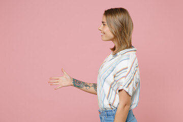 Side view of smiling young blonde woman girl in casual striped shirt posing isolated on pastel pink background. People lifestyle concept. Mock up copy space. Stand with outstretched hand for greeting.