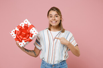 Smiling young blonde woman girl in striped shirt isolated on pastel pink background. Valentine's Day Women's Day birthday, holiday concept. Point index finger on red present box with gift ribbon bow.