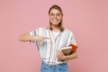 Smiling young student girl in casual striped shirt eyeglasses posing isolated on pink background. Education in high school university college concept. Mock up copy space. Point index finger on books.