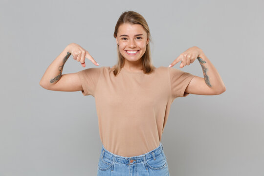 Smiling young blonde woman girl in casual beige t-shirt posing isolated on gray background studio portrait. People sincere emotions lifestyle concept. Mock up copy space. Pointing index fingers down.