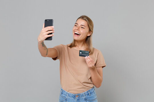 Funny young blonde woman girl in casual beige t-shirt posing isolated on gray background studio. People lifestyle concept. Mock up copy space. Doing selfie shot on mobile phone hold credit bank card.