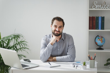 Confused young bearded business man in gray shirt sitting at desk work on laptop pc computer in light office on white wall background. Achievement business career concept. Put hand prop up on chin.