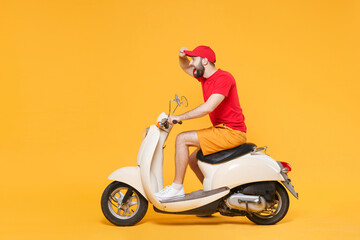 Delivery man in red cap t-shirt uniform driving moped motorbike scooter isolated on yellow background studio Guy employee working courier Service quarantine pandemic coronavirus virus covid-19 concept