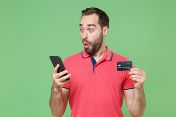 Shocked young bearded man guy in casual red pink t-shirt posing isolated on green background studio portrait. People lifestyle concept. Mock up copy space. Using mobile phone, hold credit bank card.