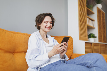Smiling young woman girl in casual clothes sit on couch spending time in living room at home. Rest relax good mood leisure lifestyle concept. Mock up copy space. Using mobile phone typing sms message.