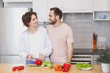 Smiling young couple friends guy girl in casual clothes preparing vegetable salad cooking food in kitchen at home. Dieting family healthy lifestyle concept. Mock up copy space. Looking at each other.