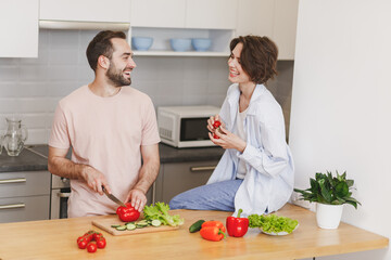 Laughing young couple two friends guy girl in casual clothes sit on table preparing vegetable salad cooking food in light kitchen at home. Dieting family healthy lifestyle concept. Mock up copy space.