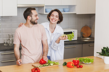 Laughing cheerful young couple two friends guy girl in casual clothes preparing vegetable salad cooking food in light kitchen at home. Dieting family healthy lifestyle concept. Mock up copy space.