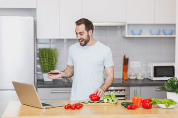 Fototapeta na wymiar Excited young man guy in casual t-shirt using laptop computer making video call preparing vegetable salad cooking food in light kitchen at home. Dieting healthy lifestyle concept. Mock up copy space.