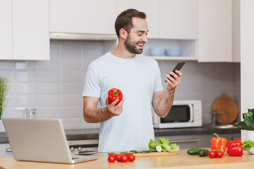 Smiling young bearded man using mobile phone looking at recipe hold bell pepper preparing vegetable...