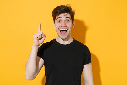 Excited young man guy 20s in casual black t-shirt posing isolated on yellow wall background studio portrait. People lifestyle concept. Mock up copy space. Holding index finger up with great new idea.