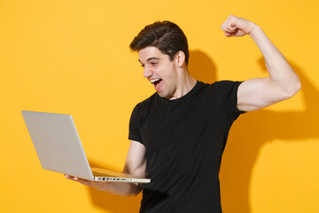 Excited young man guy in casual black t-shirt isolated on yellow background. People lifestyle concept. Mock up copy space. Working on laptop pc computer, showing biceps muscles, doing winner gesture.