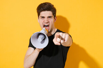 Angry young man guy in casual black t-shirt posing isolated on yellow background studio portrait. People lifestyle concept. Mock up copy space. Screaming in megaphone pointing index finger on camera.
