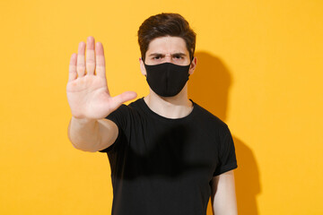 Young guy in black t-shirt face mask isolated on yellow background studio portrait. Epidemic pandemic spreading coronavirus 2019-ncov sars covid-19 flu virus concept. Showing stop gesture with palm.